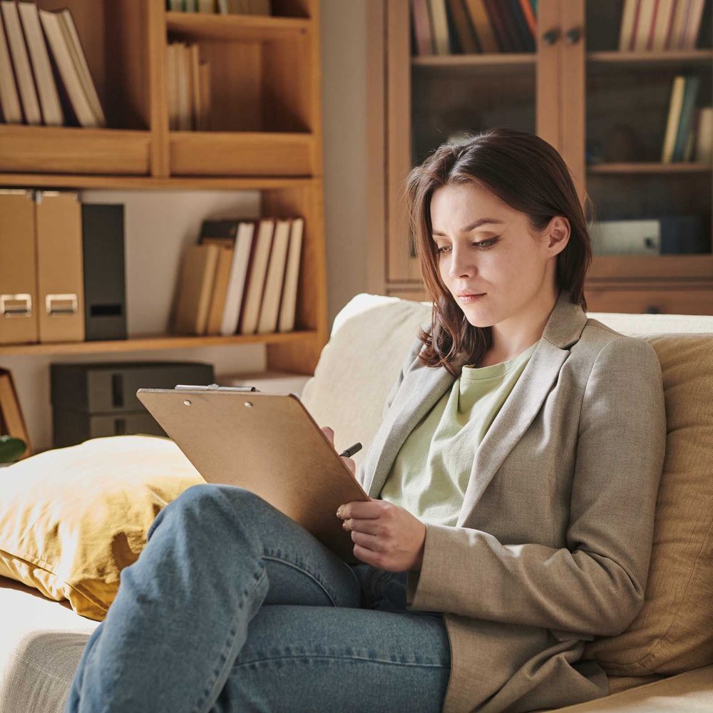 Attractive young woman sitting on sofa in modern office room holding clipboard making notes