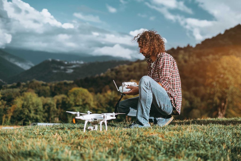 A serious bearded hipster man in a checkered shirt is sitting on the grass of a mountain meadow and preparing his remote controller to operate drone next to him; warm summer day with hills around