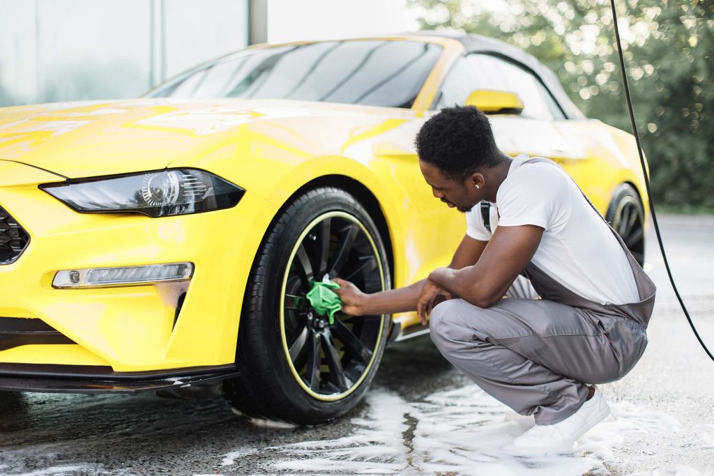 Young worker of self car wash service African bearded man cleaning a wheel, car rims of modern luxury yellow sport car with green microfiber cloth, at outdoor car wash self service