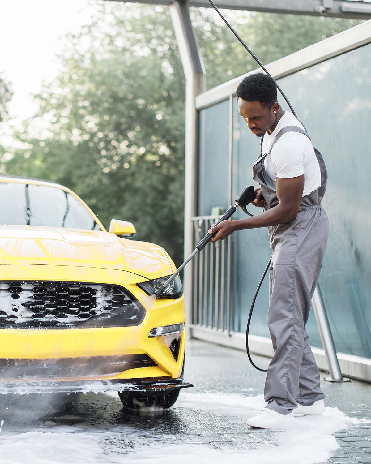 Horizontal shot of young African American man washing his luxury yellow car under high pressure water jet outdoors at self car wash station.