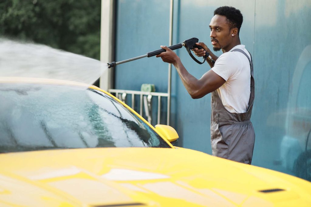 Manual car wash. Handsome African young man washing his luxury yellow vehicle with high pressure water pump at automobile cleaning self service outdoors.