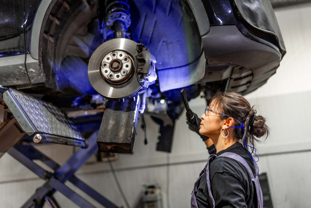 Female mechanic using flashlight, inspecting brakes of lifted car in a professional workshop, ensuring safety and performance