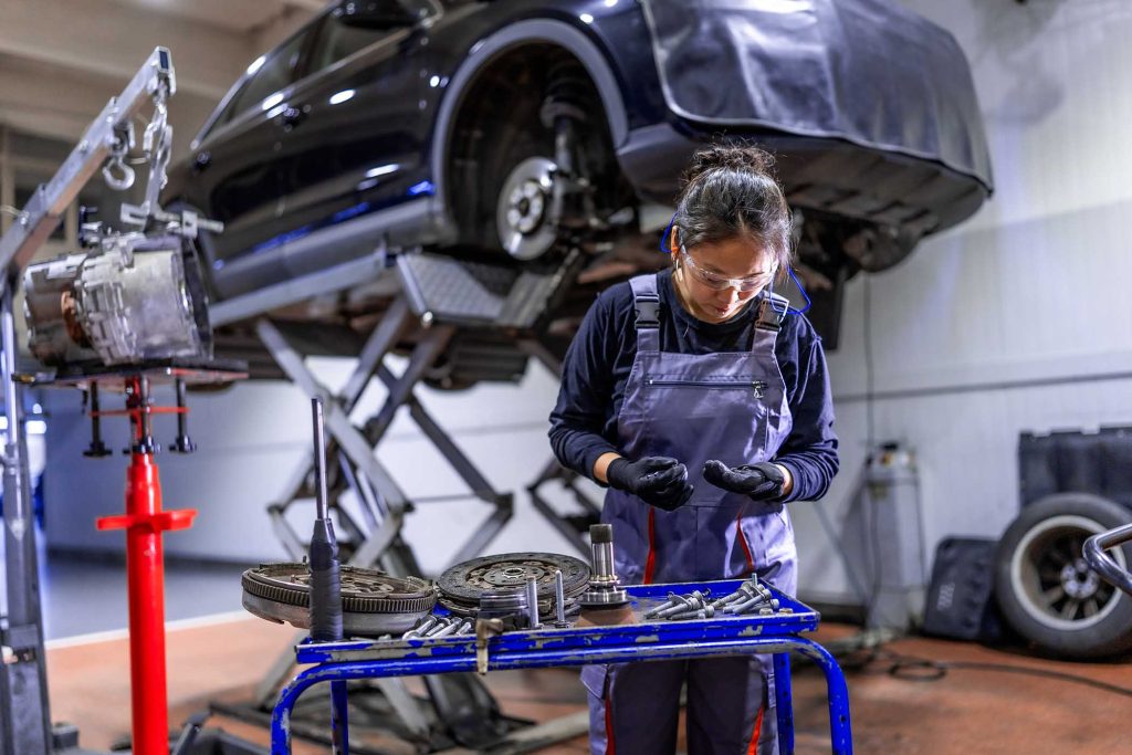 Female mechanic wearing gloves and safety glasses examining car parts with a car on a hydraulic lift in the background
