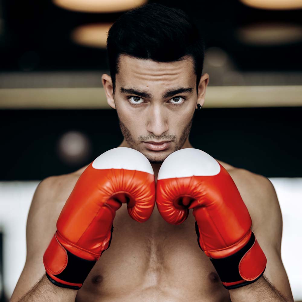 Sportsman with a naked torso keeps his hands in the red boxing gloves next to his face in the boxing gym .