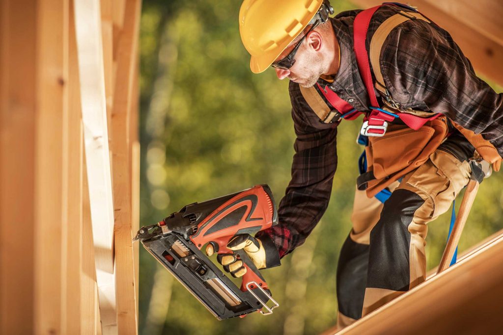 Caucasian Construction Worker with a Nail Gun Assembling Wooden Skeleton Frame Beams in a Newly Developed Building. Construction Industry Theme.