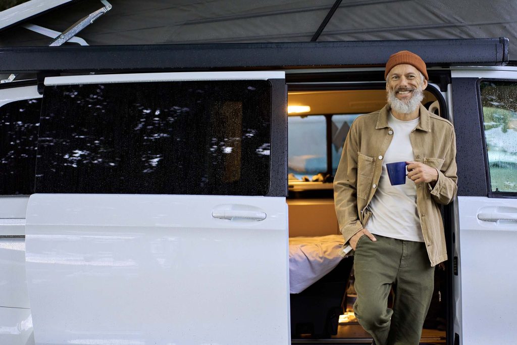 Happy older senior hipster man standing near rv camper van on vacation. Mature traveler looking at camera, holding drinking coffee waking up in the morning in camping tourism nature park.