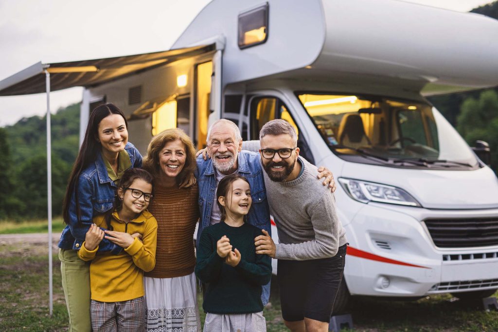 A multi-generation family looking at camera outdoors at dusk, caravan holiday trip.