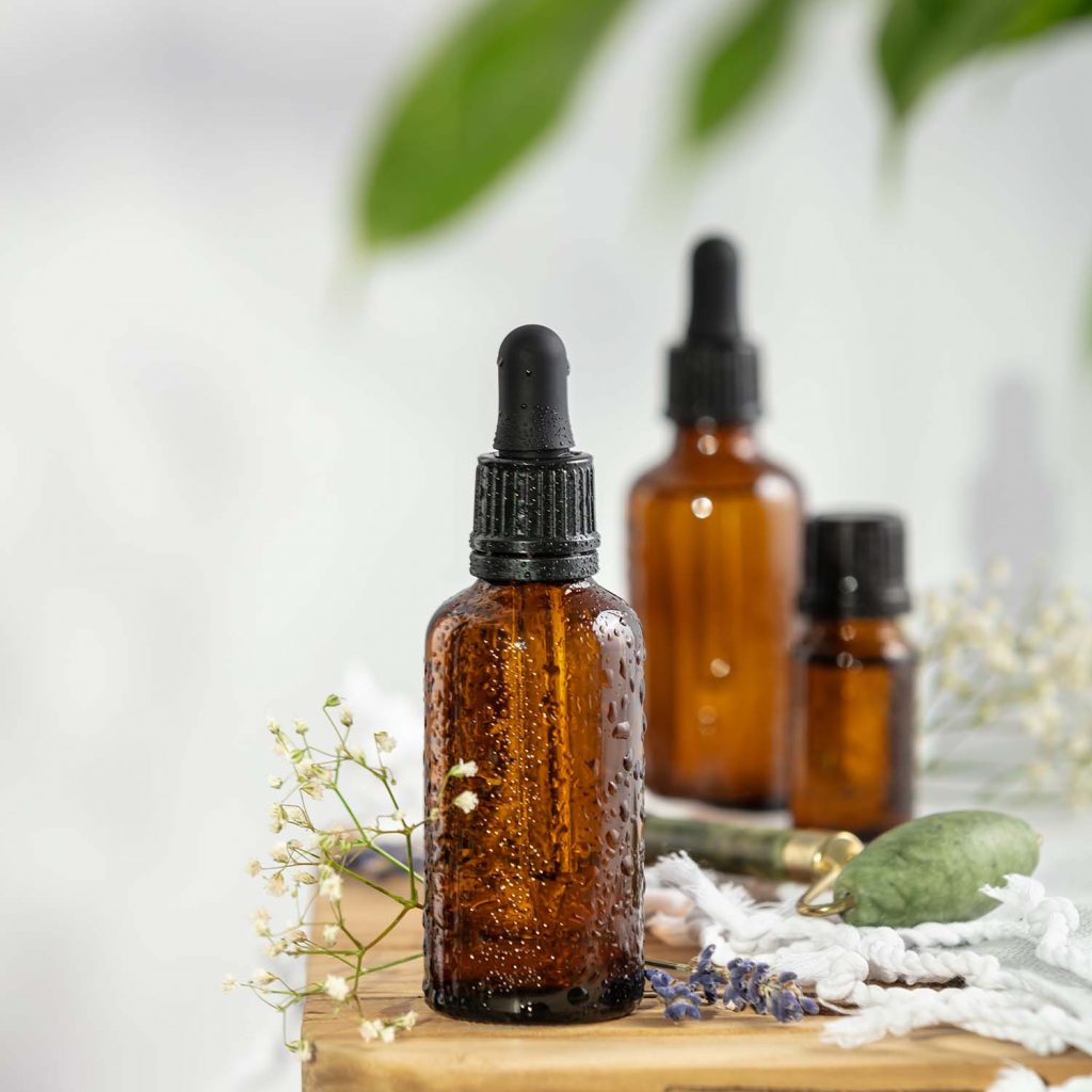 Bottles of dark amber glass with essential oil, massage jade roller and tropical leaves on wooden bench. Natural organic cosmetic, aromatherapy, message oil.
