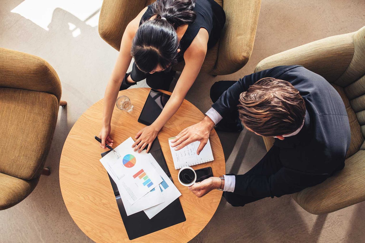 Overhead view of young businessman and woman working on graphs while sitting at office lobby. Corporate people discussing new business project using charts.