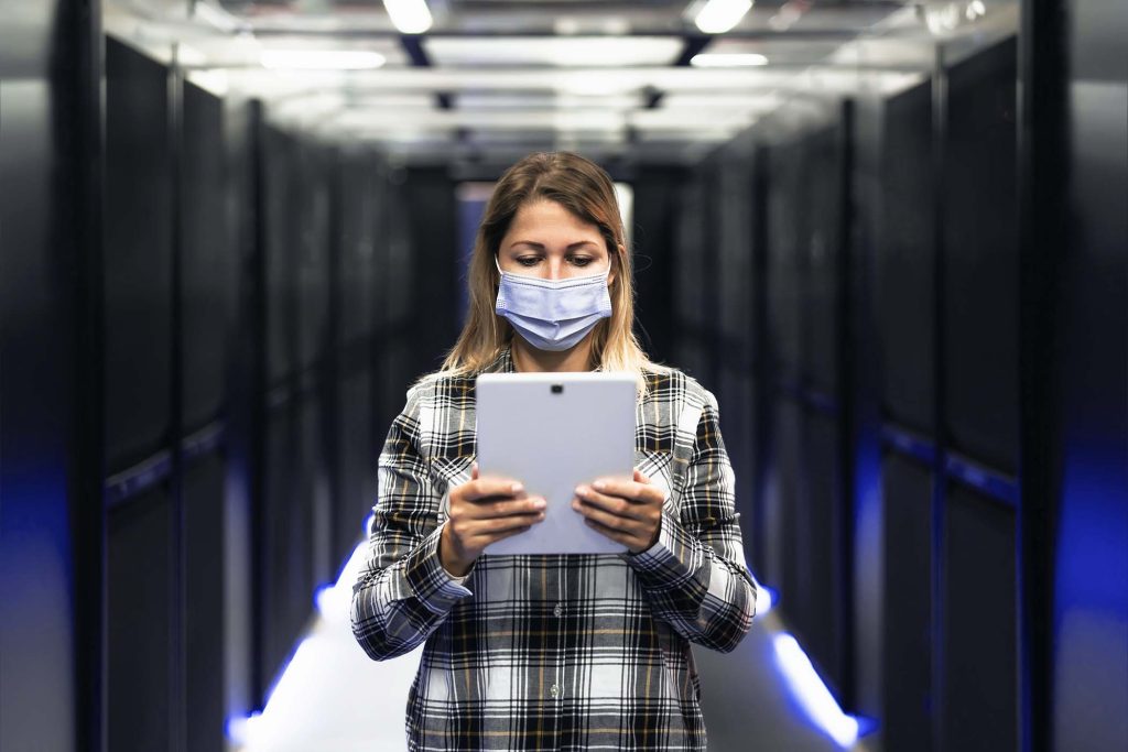 Female informatic engineer working inside server room database while wearing face mask during corona virus outbreak