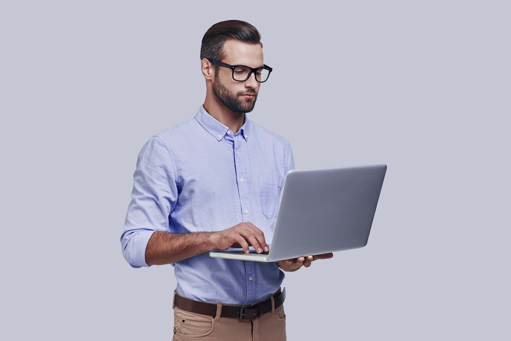 Modern technologies. Good looking young man working using laptop while standing against grey background