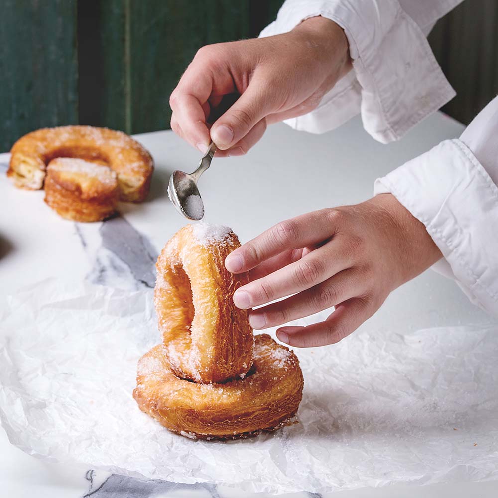 Boy's hand hold homemade puff pastry deep fried donuts or cronuts with sugar standing on crumpled paper over white marble kitchen table.