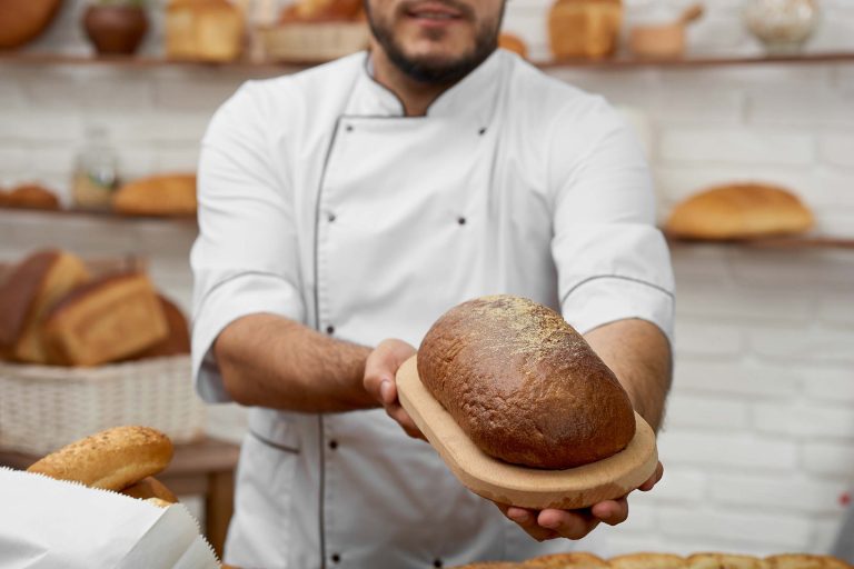 Horizontal cropped shot of a professional baker holding out loaf of delisious fresh bread posing at his store selling pastry nutrition healthy food eating organic recipe concept.