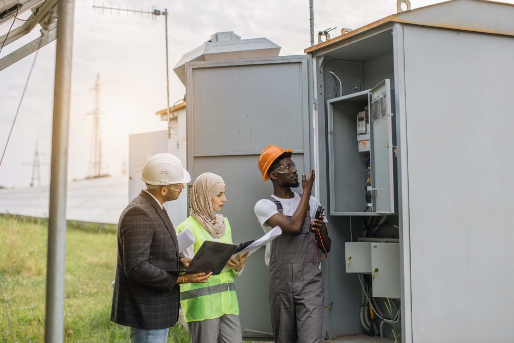 Indian man in suit and muslim woman in hijab using laptop and clipboard during examination of solar station. African american technician checking serviceability of cables in switchgear.