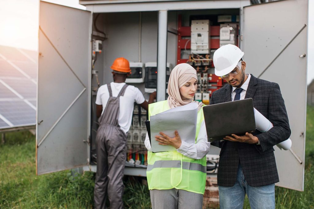 Muslim woman in hijab and indian man in suit using laptop and clipboard during discuss at solar station. African american technician working with cables in transformer on background.
