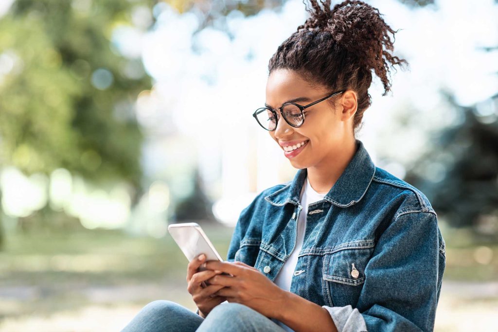 Cheerful Black Millennial Lady With Smartphone Using Mobile App Sitting In Park Outdoor. Modern Communication Concept