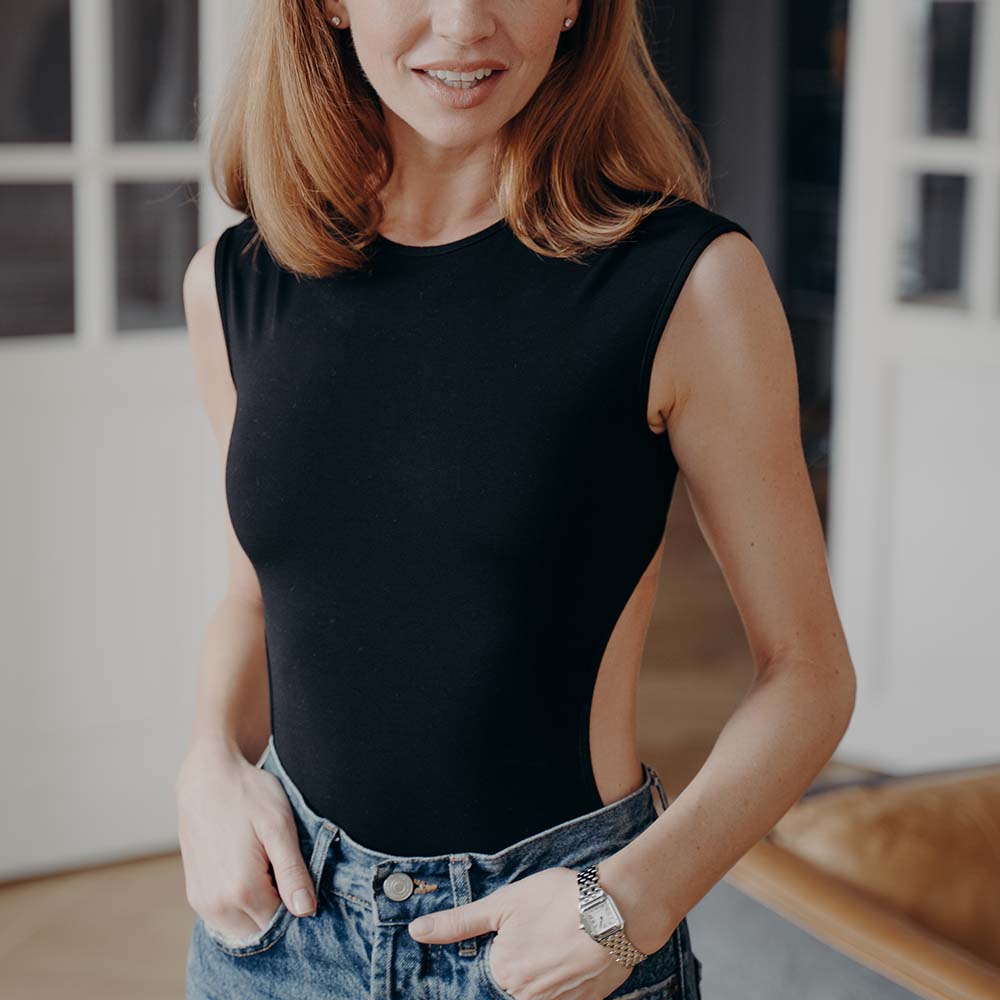 Lady in fashionable slim fit black bodysuit and jeans. Portrait of elegant blonde young woman. Redhead european model standing and smiling. Luxurious apartment interior. Concept of fashion and beauty.