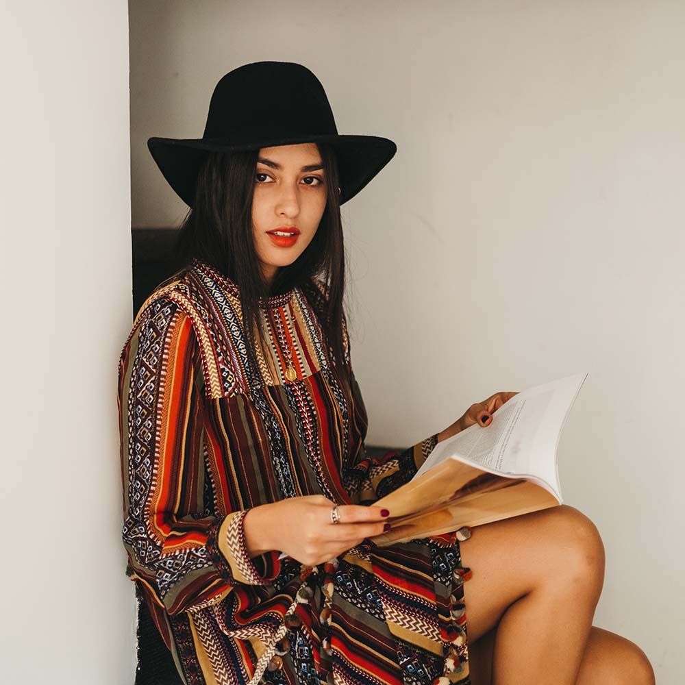 Attractive young female in stylish hat and dress looking at camera while sitting on steps and reading fashion magazine