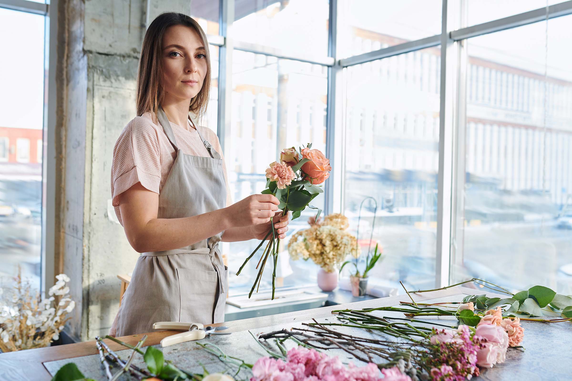 Young brunette in apron and white t-shirt looking at you while standing by table and making floral bunches in shop