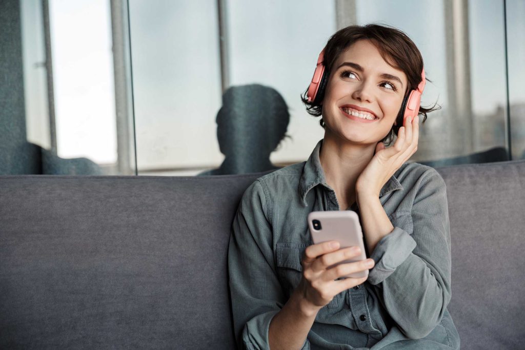 Image of nice young smiling woman using smartphone and headphones while sitting on couch at living room