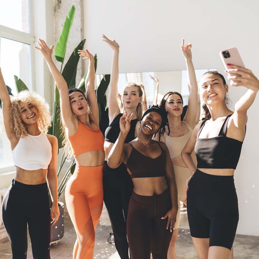 Happy multiethnic athletic women in workout tight clothes taking selfie on cellphone while standing in fitness club smiling and raising hands