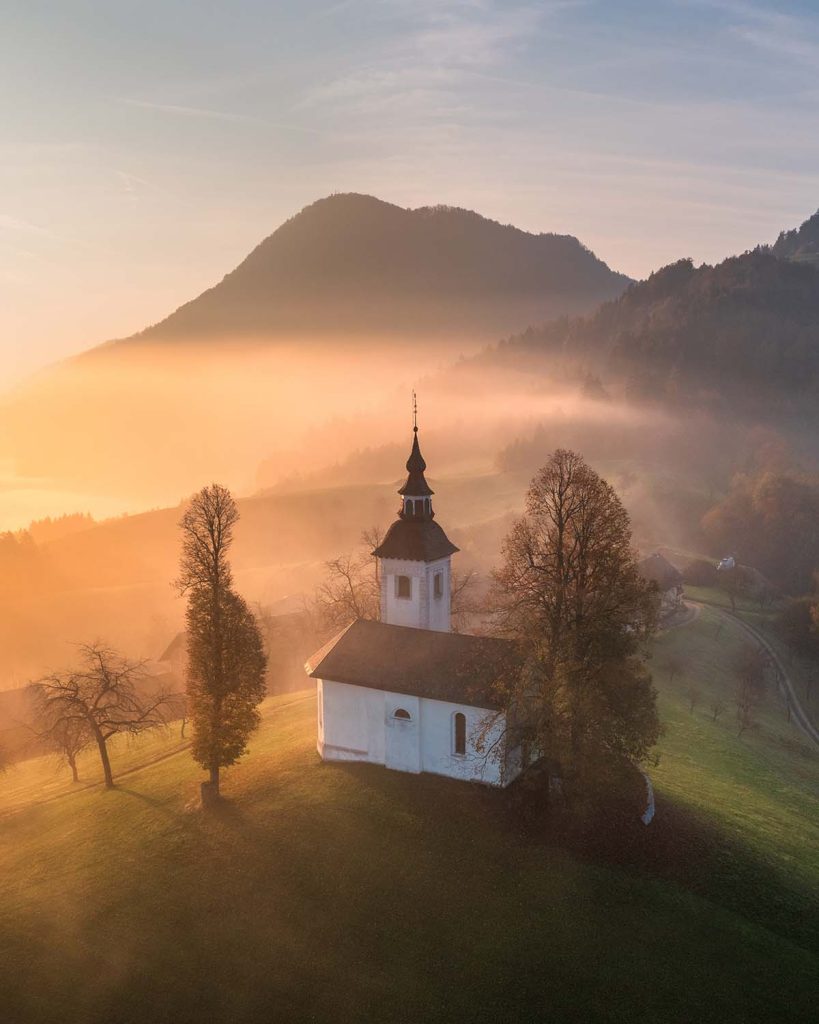Aerial view of small church on the hill in low clouds at golden sunrise in autumn. Slovenia. Top view of beautiful chapel, mountain village, fog, green meadows, trees, sky at dawn in fall. Landscape