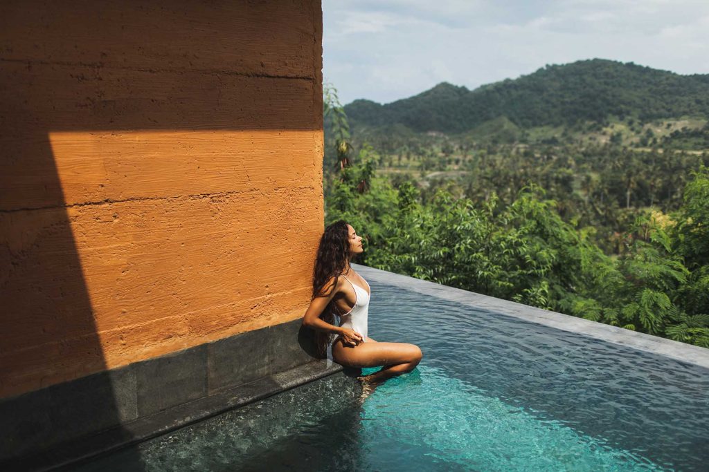 Beautiful young woman sunbathing and relaxing on edge private pool with amazing jungle and mountain view. Summer vacation, inspiration travel. White swimsuit.