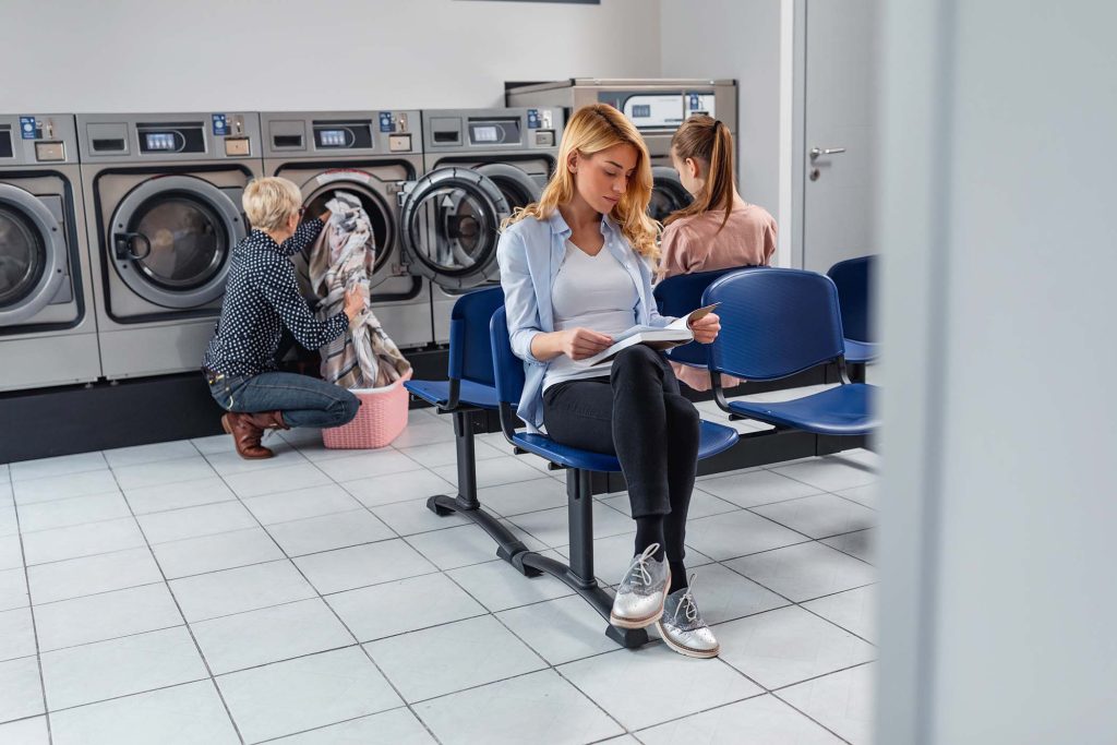 Shot of a grandmother and granddaughter using a washing machine while young woman reading a book beside