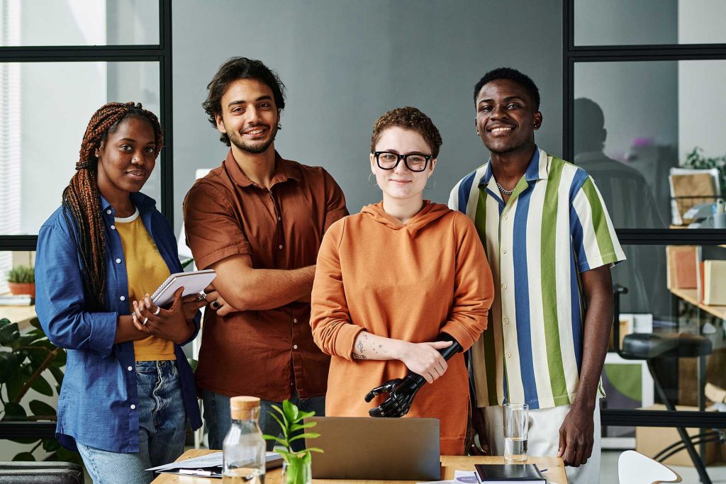 Group of young creative designers in casualwear standing by workplace with laptop and supplies in office and looking at camera