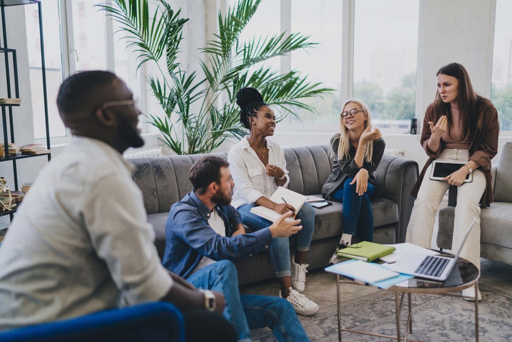 Group of diverse colleagues with documents and gadgets having discussion while sharing ideas during conversation sitting on sofa in creative workplace