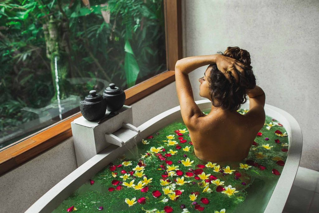 Beautiful young woman enjoying in spa, view from behind. Luxury stone bath tub with jungle view in window. Tropical flowers in water. Beauty treatment concept.