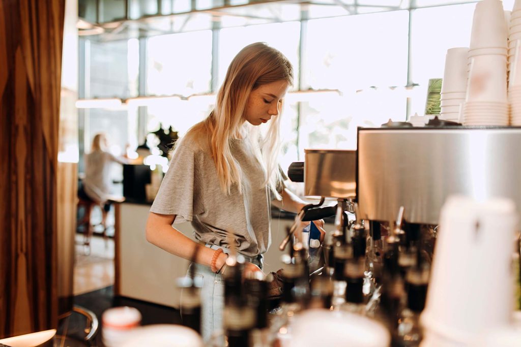 A young pretty thin blonde,dressed in casual outfit,is cooking coffee in a popular coffee shop. .