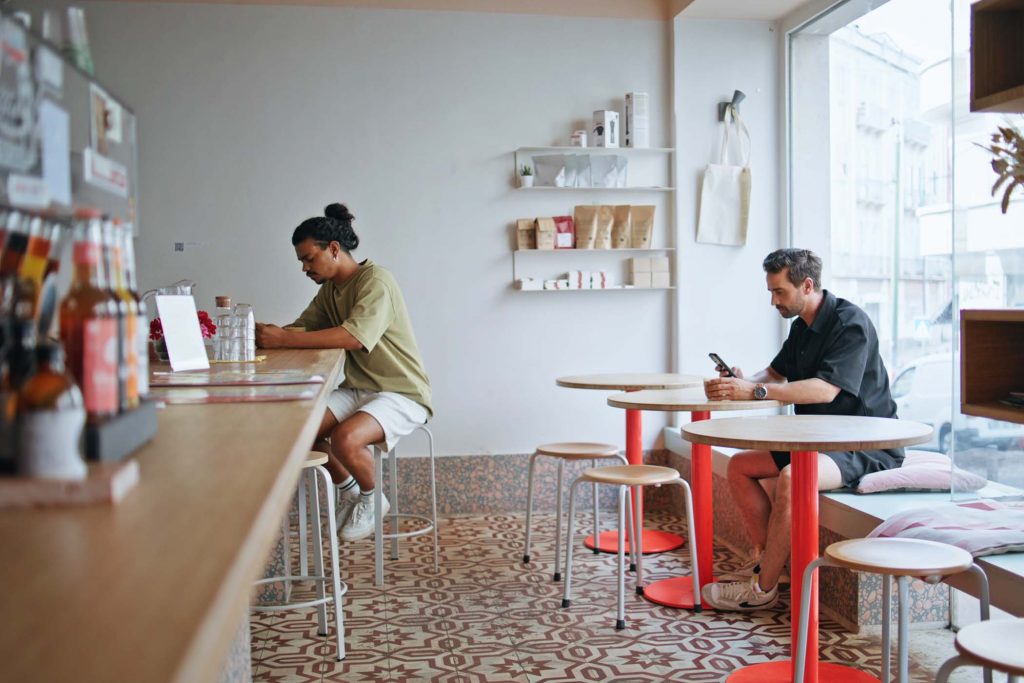 Two coffee shop clients sitting separately in cozy institution interior. Multiracial visitors cafeteria enjoying tasty caffeine drink. Couple relaxed men relaxing in coffeehouse with smartphones.