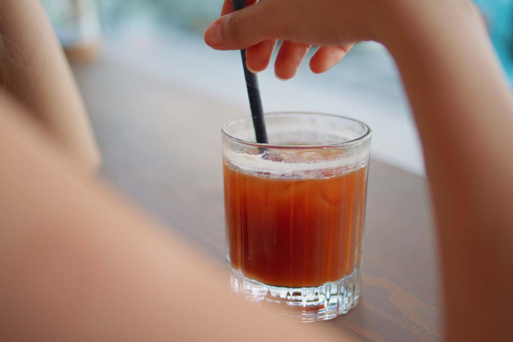 Unrecognizable woman drinking from straw cool cocktail at street cafe close up. Girl hand stirring beverage with ice cubes on wooden table. Young restaurant visitor enjoy tasty alcohol resting alone.