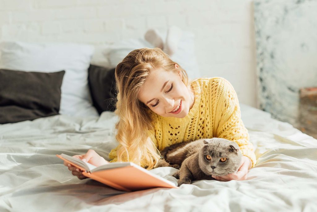 beautiful smiling young woman reading book while lying in bed with scottish fold cat