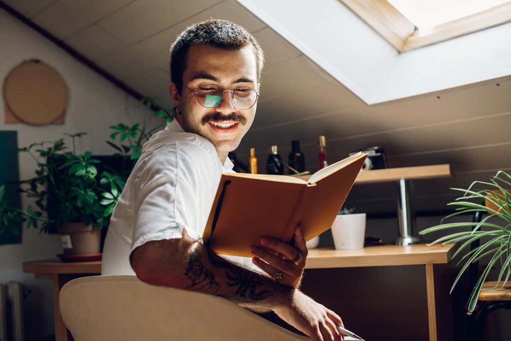 Fashionable hipster man with mustache reading a book while at home. Young man in stylish clothes relaxing inside his cozy loft. Male enjoying reading interesting book while sitting on a chair.