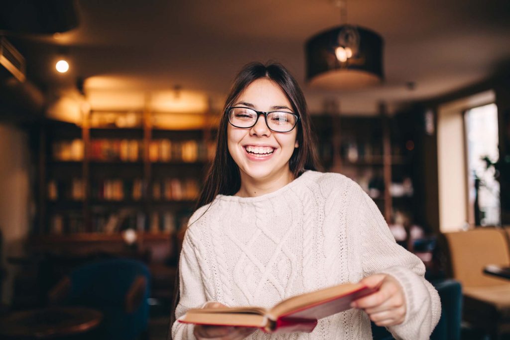 Smiling young female student laughing and holding a book in library