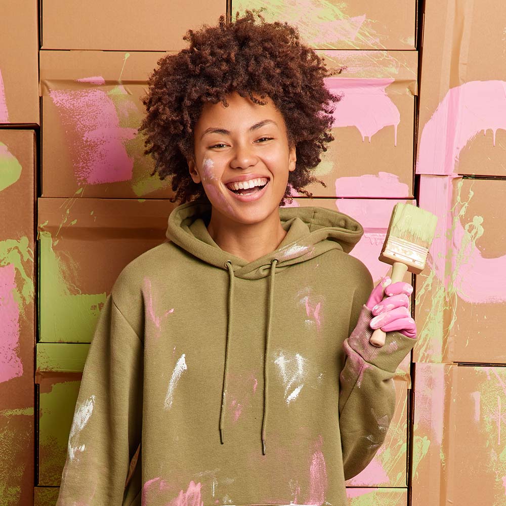 Positive ethnic woman does renovation at home holds bucket with paint and brush has happy expression as almost finished work refurbishes walls of new apartment dressed in casual dirty clothes