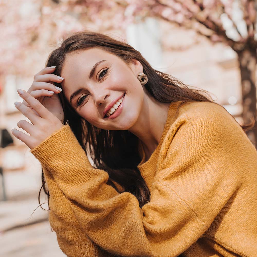 Beautiful woman in bright sweater posing on street against background of sakura. City portrait of attractive lady in yellow outfit smiling widely