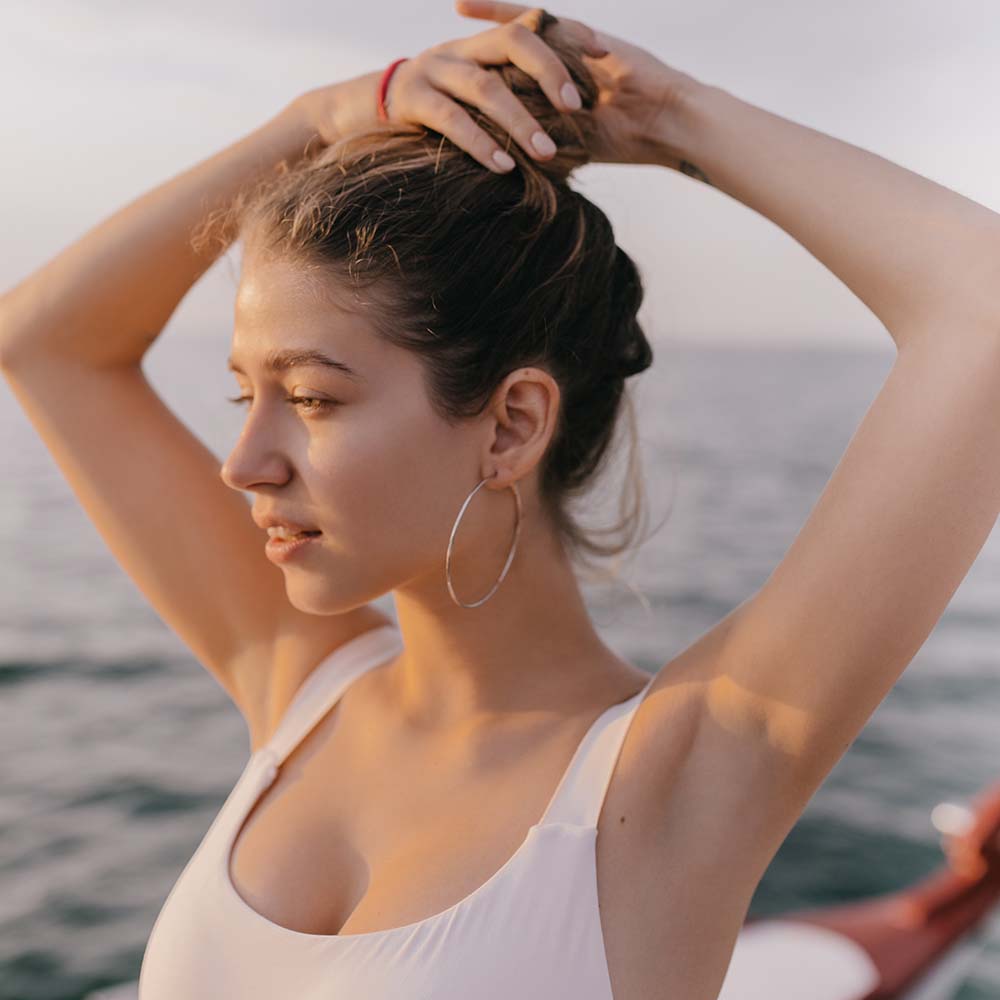 Tanned young lady in white swimsuit and stylish round earrings gathering light hair in elastic band against background of blue sky and sea