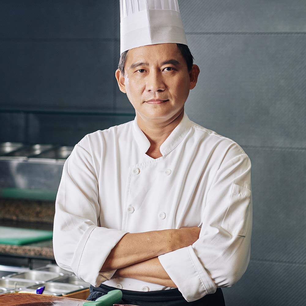Portrait of Asian mature chef in uniform standing with arms crossed near the kitchen table and looking at camera