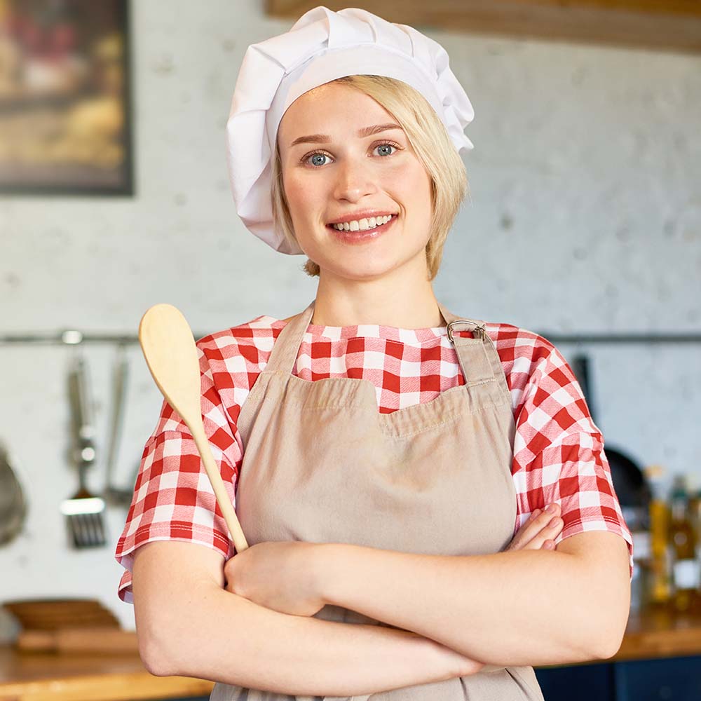 Waist-up portrait of pretty young woman wearing apron and chefs hat posing for photography with toothy smile while standing at kitchen table with wooden spoon in hands.