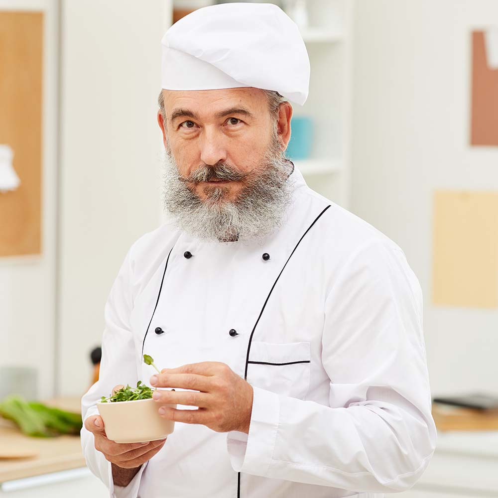 Waist up portrait of bearded senior chef looking at camera holding bowl with fresh herbs while standing in restaurant kitchen, copy space
