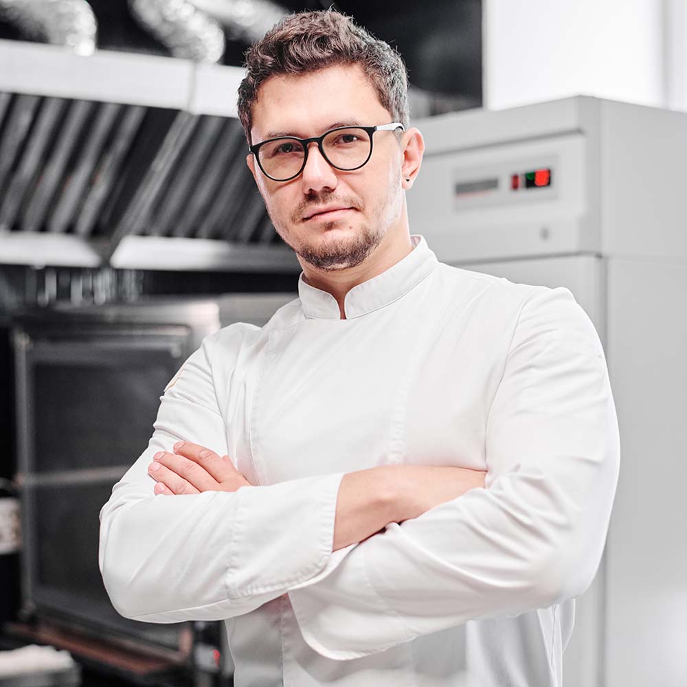 Portrait of young chef in eyeglasses and white uniform standing with his arms crossed in kitchen and looking at camera
