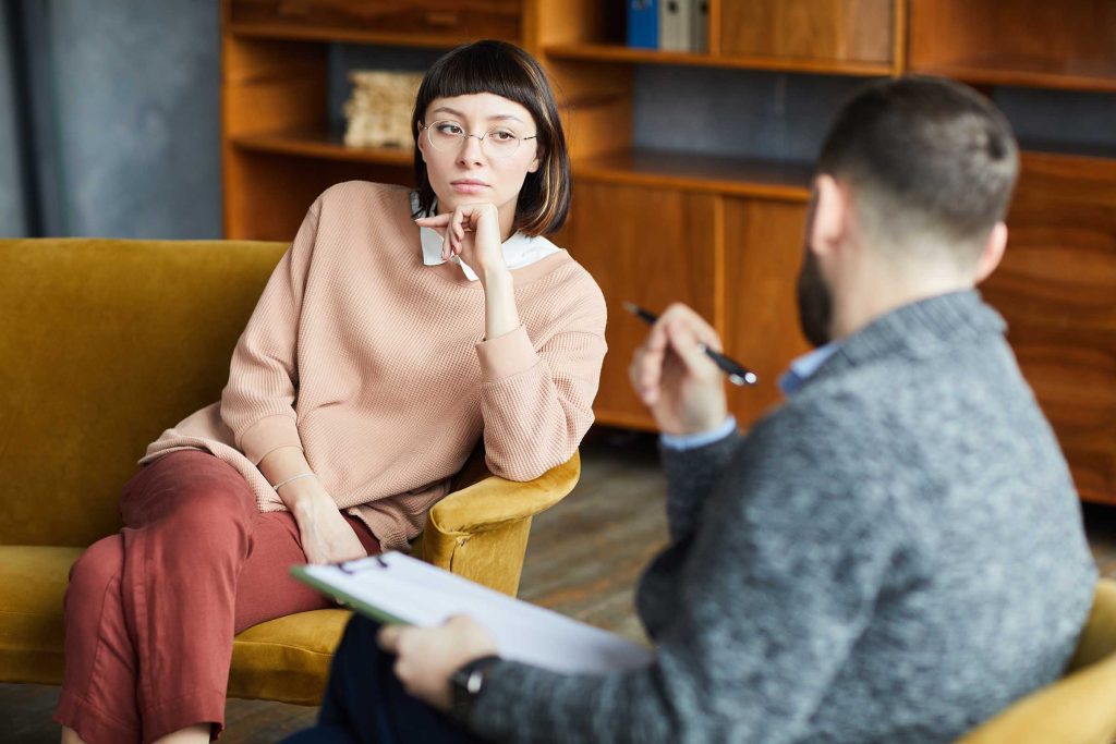 Young woman in eyeglasses sitting on sofa and talking to the man during her visit of psychology therapy