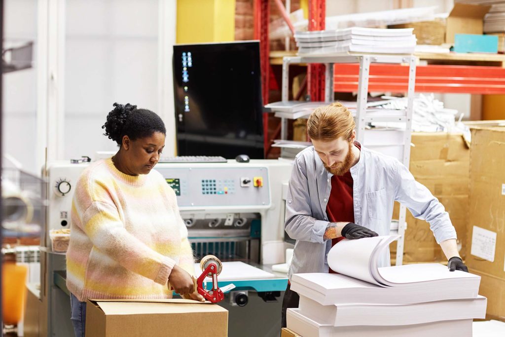 Portrait of two workers opening boxes with paper in industrial print shop, copy space