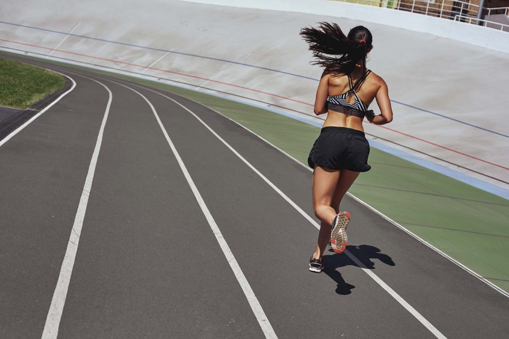 Young woman wearing black sportswear running on racetrack during training session. Female runner practicing on athletics race track. Back view.