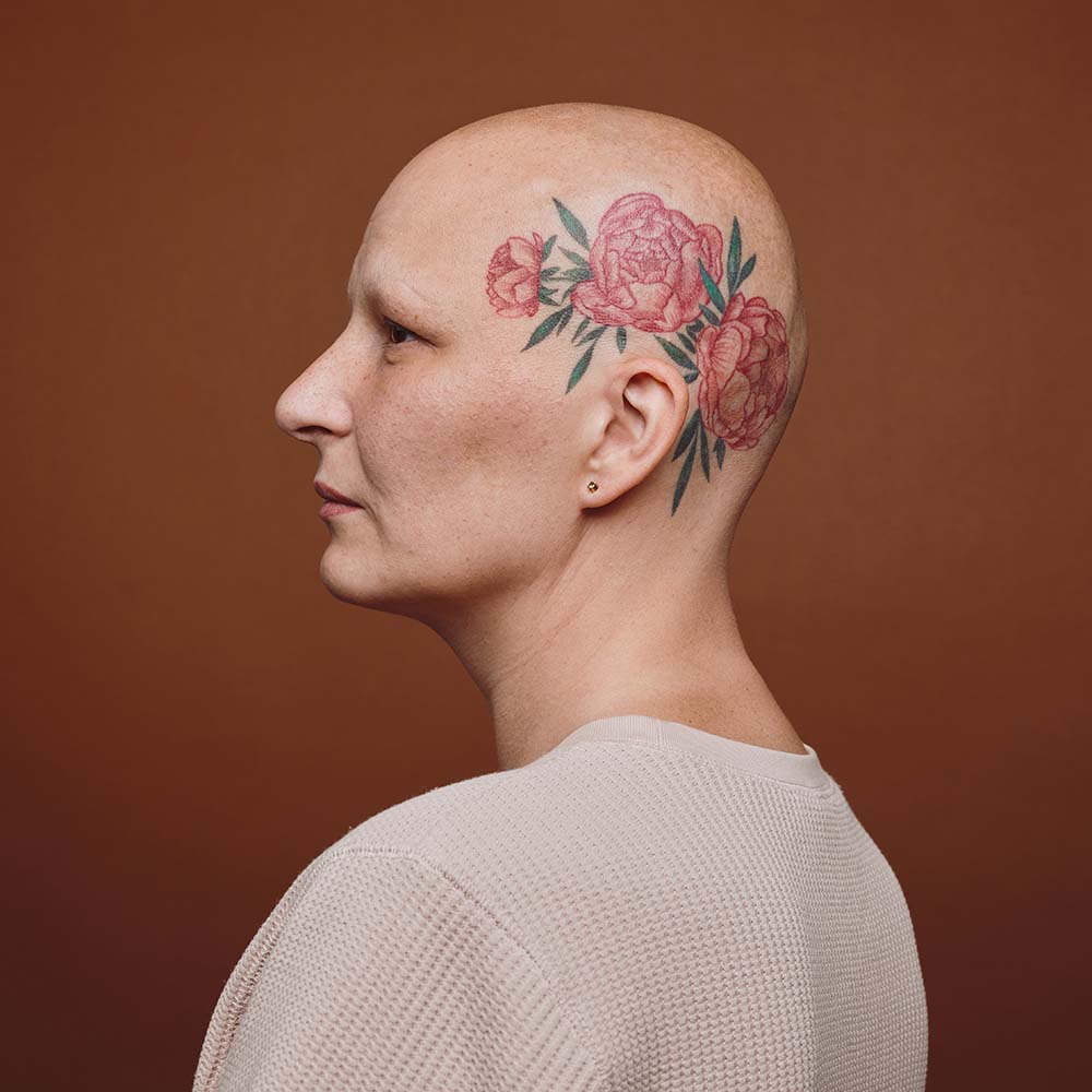 Minimal side view portrait of confident bald woman with head tattoo posing against brown background in studio, alopecia and cancer awareness, copy space