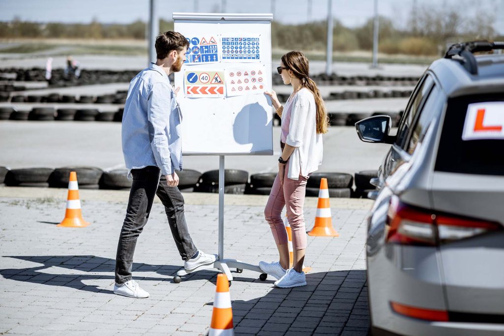 Male instructor showing traffic signs to a young female student standing on the training ground at the driver's school