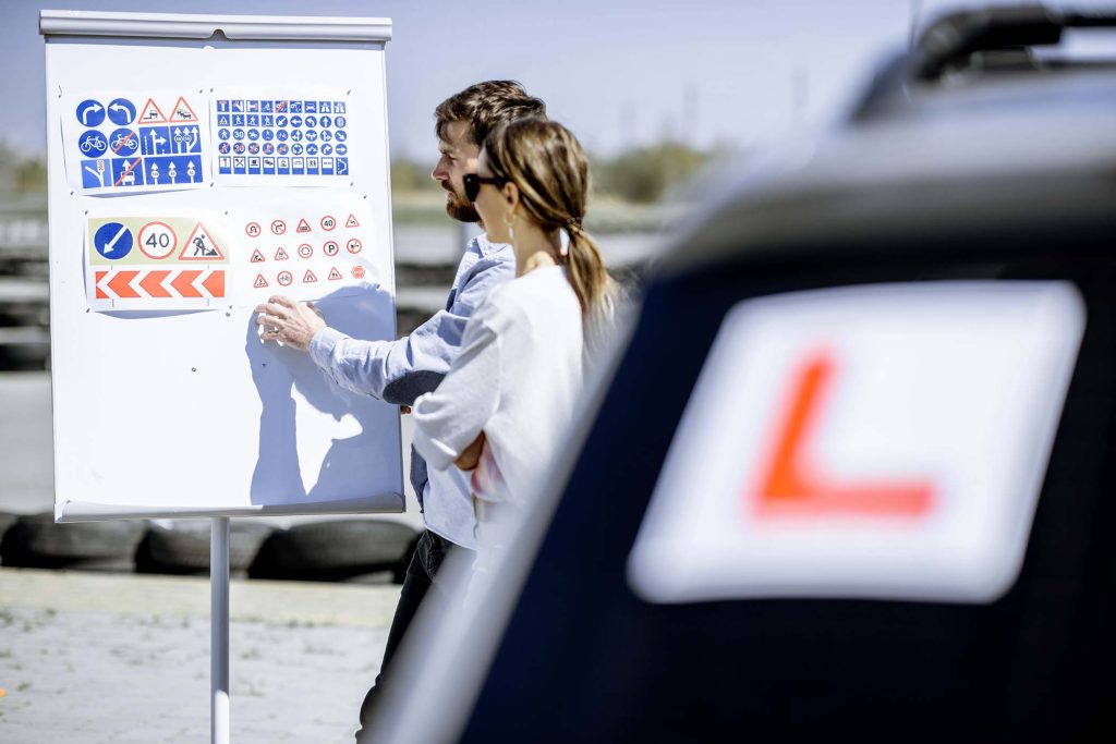 Male instructor showing traffic signs to a young female student standing on the training ground at the driver's school